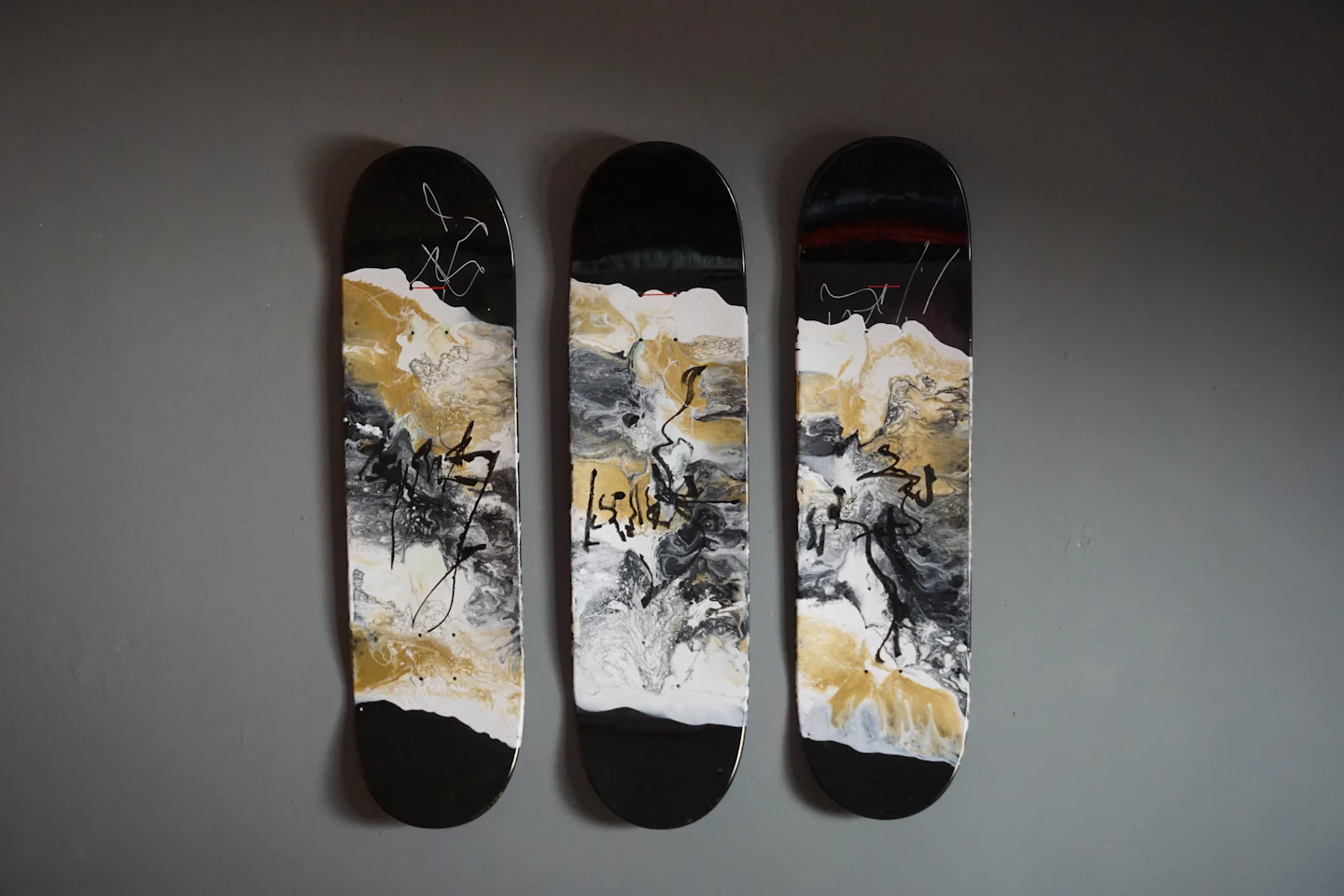 Trois planches de skate aux motifs abstraits noirs, blancs et dorés, accrochées à la verticale sur un mur gris